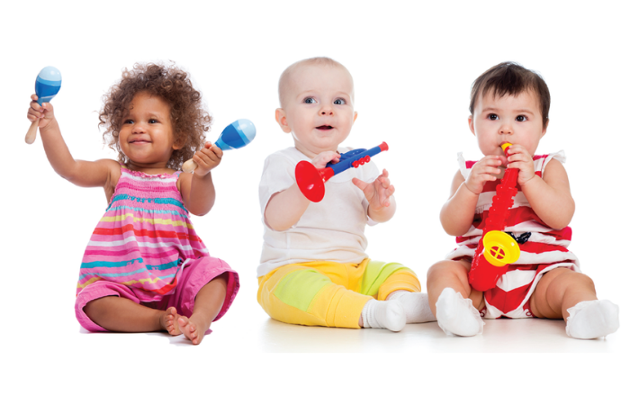 3 babies or toddlers in a row in bright clothes. From left to right - a little Black girl in a pink dress with maracas, a little white baby in white and yellow with a trumpet and a little white  baby in red and white with a saxophone toy