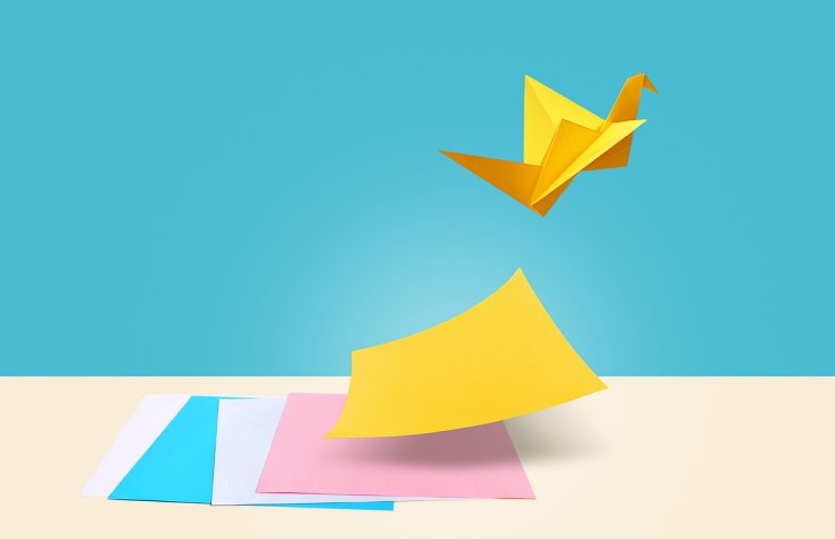 A few coloured square papers at table. Top sheet almost floating up and another is made up into a swan shape and is flying off.